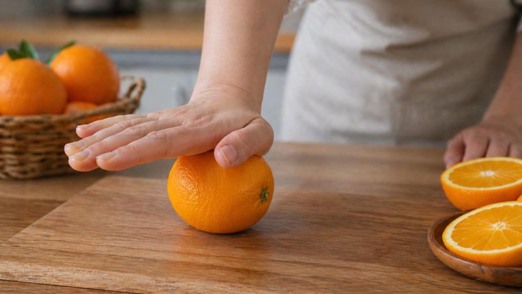Hình ảnh tay lăn nhẹ quả cam trên mặt bàn để làm mềm múi cam, giúp vắt dễ hơn và hạn chế vị đắng