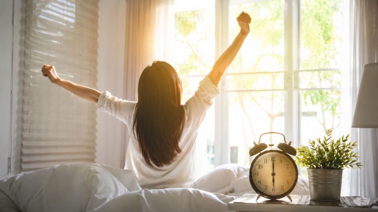 Một người thức dậy sớm đầy năng lượng vào buổi sáng với ánh nắng tự nhiên trong phòng ngủ - Mẹo dậy sớm mà không mệt mỏi hiệu quả mỗi ngày